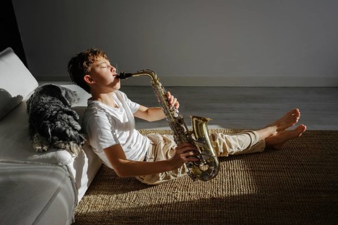 A boy sits on the floor, playing a saxophone in a sun lit room. His dog relaxes nearby on the couch