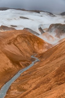 Steaming stream between colourful rhyolite mountains with snowfields, Hveradalir geothermal area,