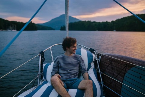 A man relaxes on a sailboat, enjoying the tranquil afternoon. The serene water and distant