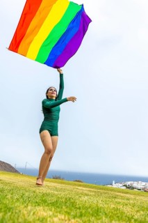 A joyful woman dances outdoors, waving a colorful LGBTQIA+ pride flag. Her lively expression and
