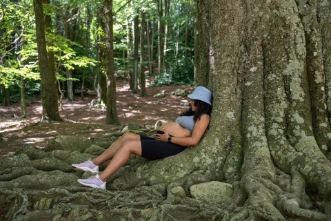 A pregnant latin woman wearing a hat sits peacefully against a large tree in a serene forest,
