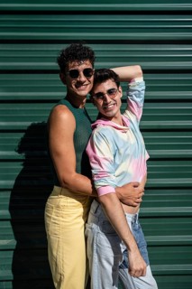 A cheerful gay couple sharing an affectionate hug, standing by a green wall on a sunny day. Both