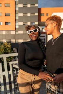 A cheerful LGBT multiethnic couple stands on a balcony, basking in the sunlight. They wear trendy
