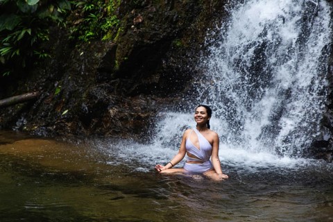 A young Ecuadorian woman joyfully sits in a natural pool, with a lush waterfall in the background