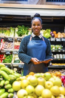 Smiling saleswoman wearing apron holding tablet in organic supermarket with fresh fruits and