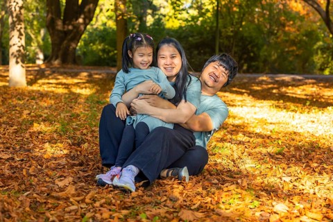 Asian family of three, smiling and embracing together, sitting on a bed of autumn leaves in a
