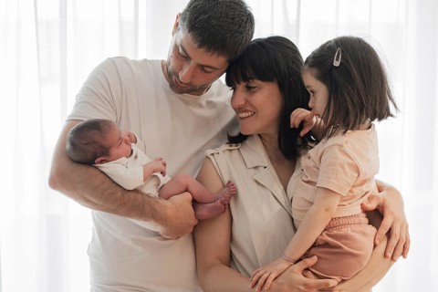 A joyful young family embraces in a bright room, with a proud father holding a newborn baby and a