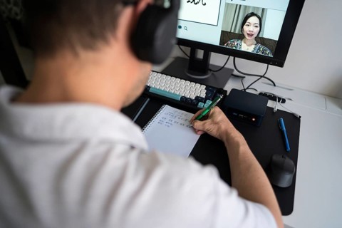 A man is focused on learning Mandarin through an online class at home. He writes characters in a