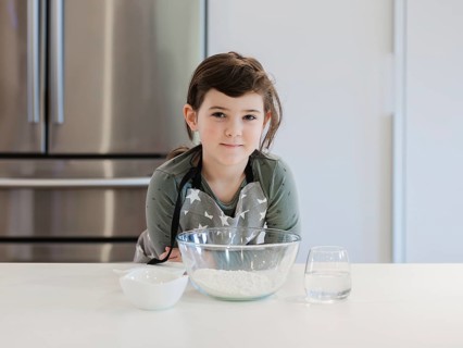 A young child, aspiring chef stands confidently at a kitchen counter with baking ingredients ready.