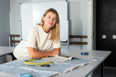 A woman leans over a desk in a sewing studio, surrounded by fabric, scissors, and a measuring tape,