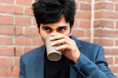 A indian businessman in a blue suit enjoys a coffee break, sipping from a cup. He stands against a