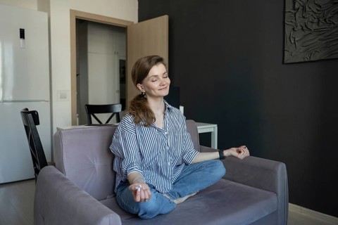 A woman sits cross-legged on a sofa, meditating with closed eyes in a serene home environment. She