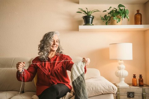 A smiling woman sits on a comfortable sofa, knitting a gray garment in a warmly lit living room.