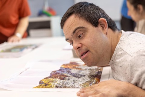 A man with Down syndrome focuses intently on a colorful mosaic artwork, carefully placing beads