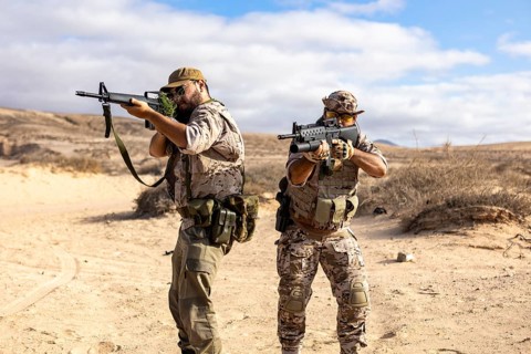 Two soldiers in tactical gear engage in a military exercise in a desert landscape, aiming their