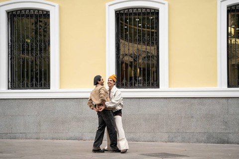 A Colombian gay couple joyfully embraces in front of a yellow building with arched windows, sharing