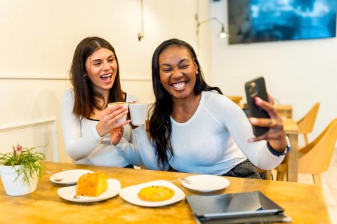Two happy female friends are making a selfie while toasting with cappuccino cups and having