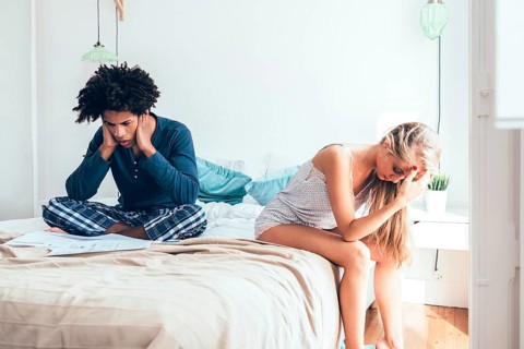 A distressed couple sits on a bed, each showing signs of stress. They are surrounded by papers,