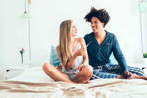 A couple sits on a bed, joyfully reacting to positive pregnancy test results. The room is bright,