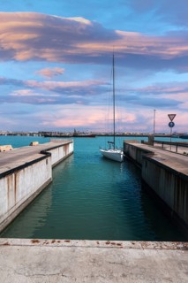 A sailboat calmly docked in a marina canal in Civitanova Marche, Italy. The sky is painted with
