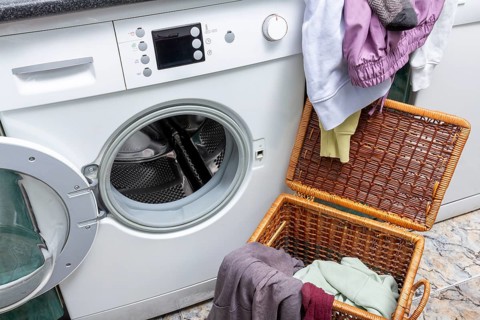 A laundry basket full of colorful clothes sits in front of an open front-loading washing machine,