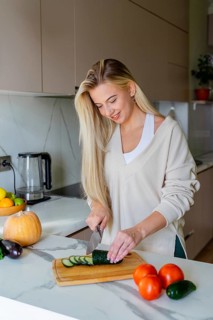 A woman in a cozy kitchen slices cucumbers on a wooden board. Surrounded by fresh vegetables, the