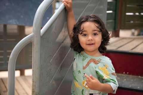 A toddler with curly hair smiles happily while standing next to a slide in a sunny park. He is