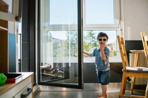 A cheerful boy with Down syndrome, wearing sunglasses, stands by an open glass door, enjoying the