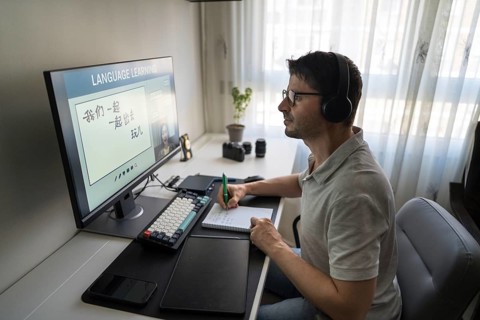 A man sits at his desk, wearing headphones, as he studies Mandarin online via a computer. The
