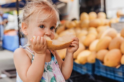 A joyful toddler savoring a fresh slice of melon at a bustling local bazaar. The vibrant market