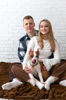 A cheerful young couple is sitting on the floor at home, lovingly embracing their playful white and