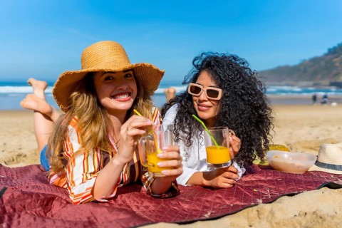 Two smiling women are relaxing on a blanket on the sand, holding glasses of refreshing orange juice