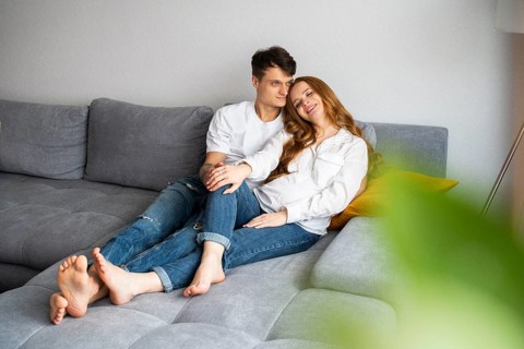 A young couple, expecting a baby, sits lovingly on a cozy gray couch, embracing and smiling warmly.