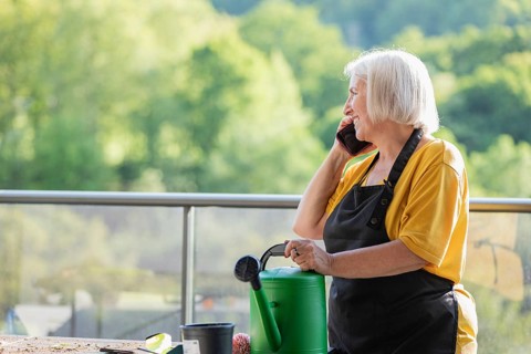 An elderly woman in a yellow shirt and black apron talks on the phone while gardening on a balcony.