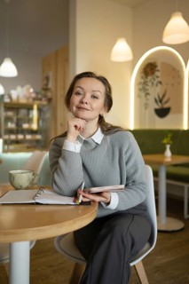 A woman sits at a cafe table, pensively holding a tablet and pen, with an open notebook and coffee