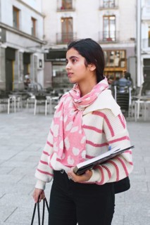 A arab student woman stands in a city street holding a notebook and tote bag. She wearing a pink
