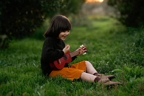 A joyful child sits on lush grass with orange trees at sunset, playing a red ukulele. The scene