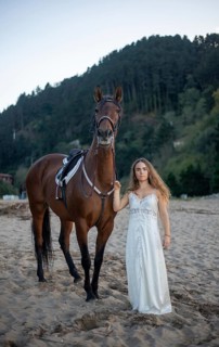 A young woman in an elegant white dress stands beside a majestic horse on a sandy beach. The serene