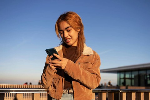 A Mixed-race woman stands outdoors using her smartphone, wearing a brown jacket and smiling The sky