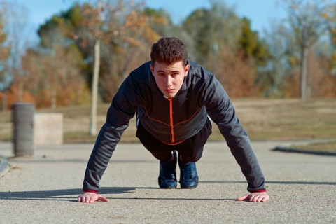 A young man performs push ups on a concrete path in a park. He is focused and dressed in sporty