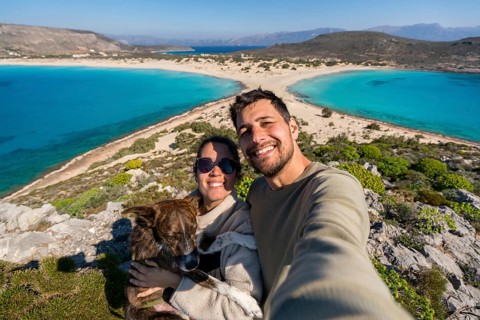 A joyful couple takes a selfie with their dog against the backdrop of a stunning, turquoise beach