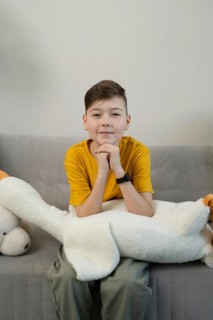 A boy wearing a yellow shirt sits on a gray sofa, resting his arms on a large white plush toy. The