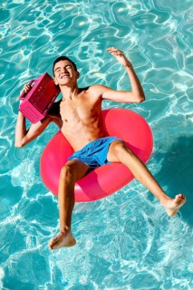 A teenage boy relaxes on a pink inflatable ring in a clear pool, holding a boombox and smiling