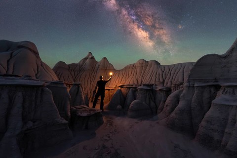 A lone figure holding a light stands amidst Goblin Valley's unique sandstone formations, beneath a