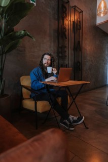 A man relaxes with a coffee in a cozy cafe, sitting with a laptop and looking contemplative. The