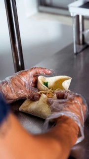 Gloved hands skillfully assemble a tasty wrap on a stainless steel surface. This scene captures the