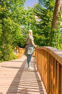 Woman walking on a sunny wooden footbridge, surrounded by green trees, Panorama Holzsteg, Calw,