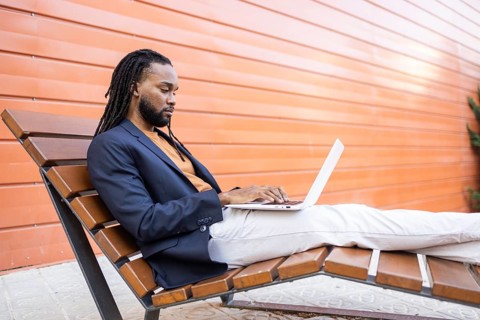 A cuban man seated on a modern outdoor bench, focused on a laptop. The orange background adds a