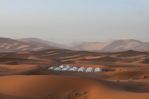 A remote Merzouga desert camp nestled among vast, rolling sand dunes under a clear blue sky. The