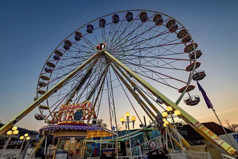 An illuminated Ferris wheel rises into the evening sky at a fair, carousel and the hustle and